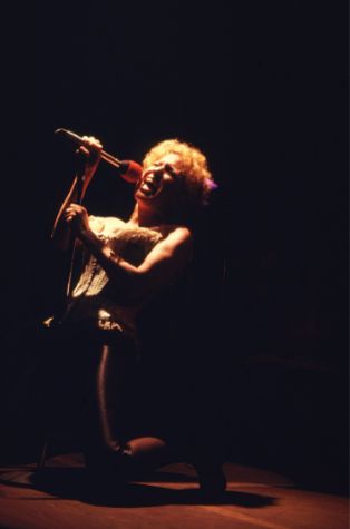 Female singer kneels on stage, singing into a microphone under dramatic spotlight.