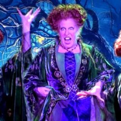 Three witches with bright red curls in green cloaks perform on a dark, forest-themed stage, arms raised and snarling under neon Halloween lighting.