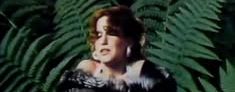 Woman in a fur stole stands among large green ferns, looking to the side.