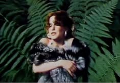 Woman in a fur stole stands among large green ferns, looking to the side.