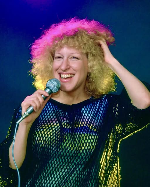Woman with curly blond hair singing into a microphone on stage, wearing a dark fishnet top with colorful lighting behind her.