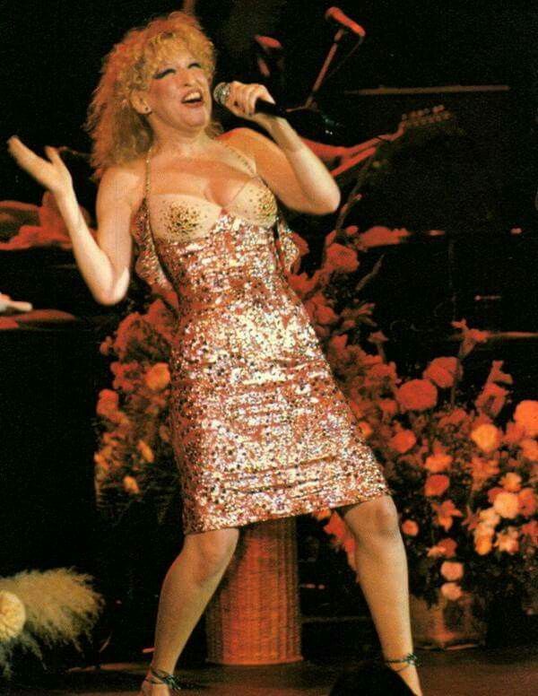 Female singer performing on stage in a sparkling pink dress, holding a microphone and mid-song gesture; floral stage backdrop behind her.