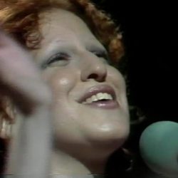 Close-up of a woman singing into a mint-green microphone on stage, smiling warmly at the crowd.