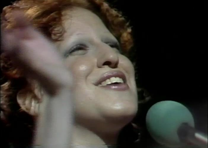 Close-up of a woman singing into a mint-green microphone on stage, smiling warmly at the crowd.