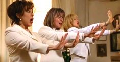 Three women in cream blouses sing with arms extended forward on a warmly lit stage, performing together.