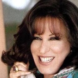 6b5652fd896827f8ff9193c95d9da647 - bootleg betty Smiling woman on a landline phone wearing a turquoise patterned blouse and gold jewelry lined with rings and bracelets.