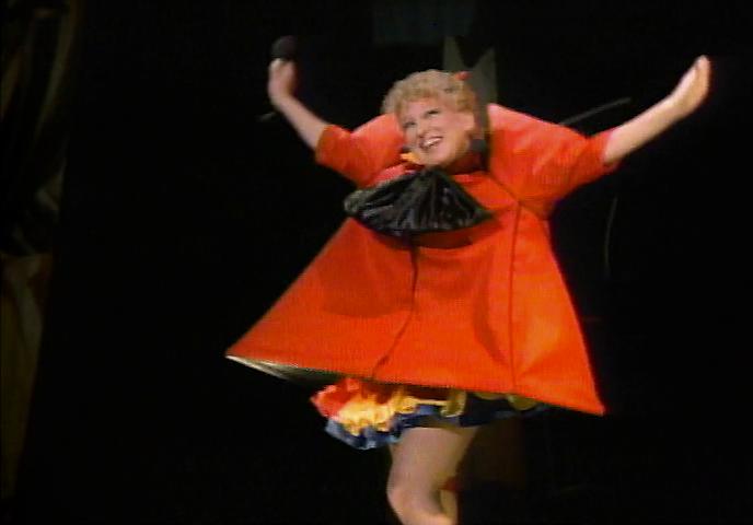 Performer in a bright red costume with arms raised on stage, smiling.