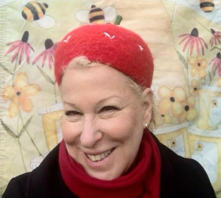 Smiling woman wearing a red hat with bee decoration and a red scarf, against a floral backdrop.