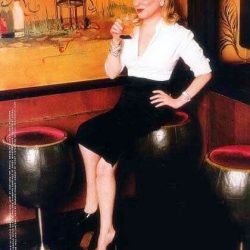 Woman in a white blouse and black pencil skirt sits on a bar stool, holding a glass in a dim, lounge-style setting with an orange mural backdrop.