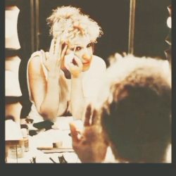 E3da2b19ba9d571ac576e200f3e23d1f - bootleg betty Person with light, curly hair applying makeup at a dressing table, mirror in front, cosmetics on the table behind them behind-the-scenes backstage scene.