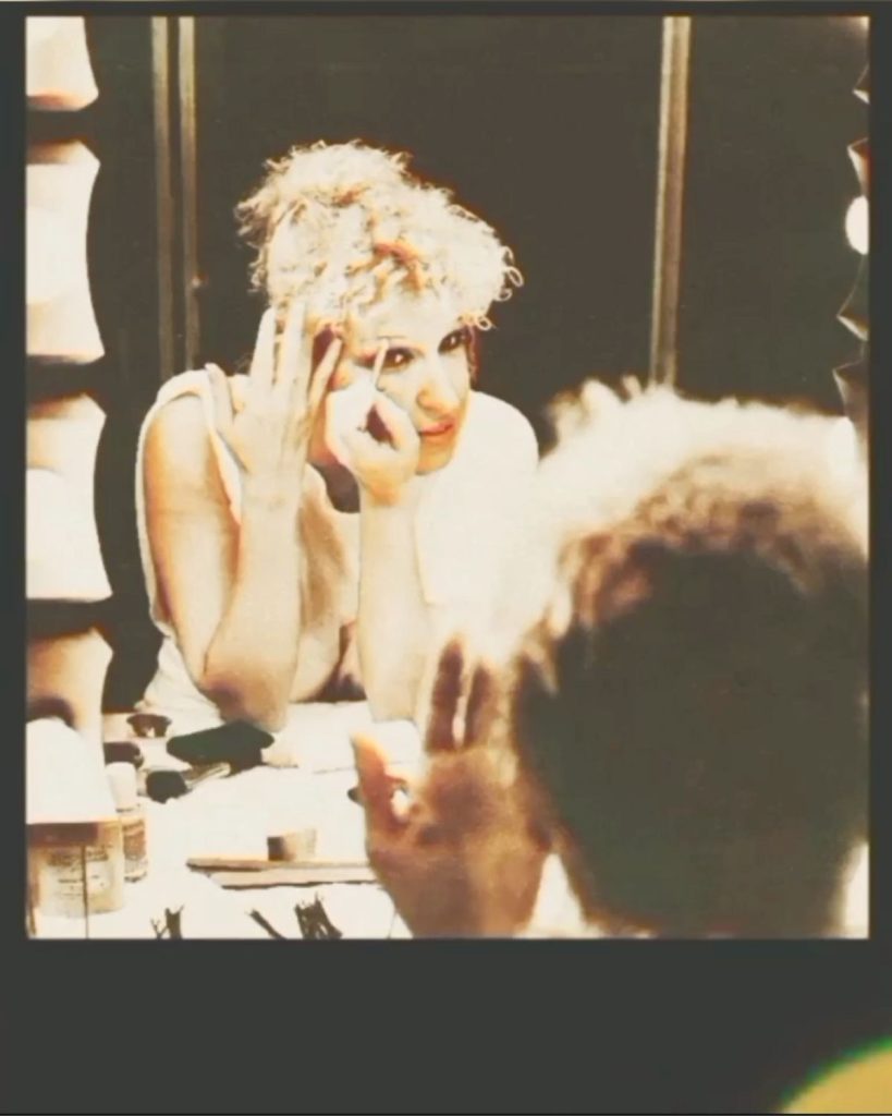 Person with light, curly hair applying makeup at a dressing table, mirror in front, cosmetics on the table behind them behind-the-scenes backstage scene.