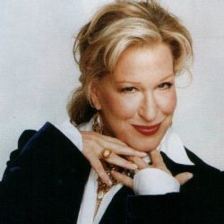 Portrait of a stylish woman with blonde hair, wearing a black blazer and white blouse, smiling with her hands near her chin.