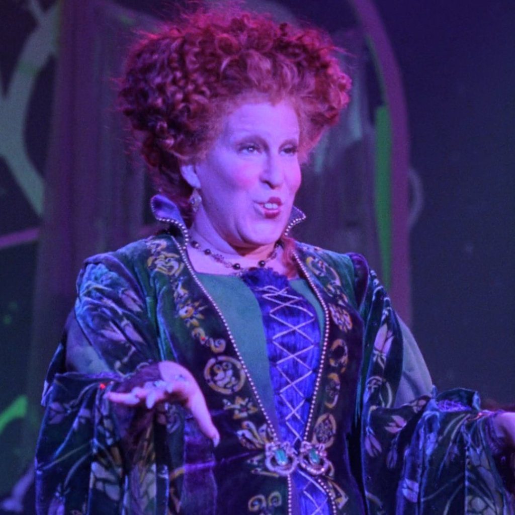 Stage performer in a green, velvet Renaissance-style dress with a purple corset, under purple lighting, speaking or singing on stage.