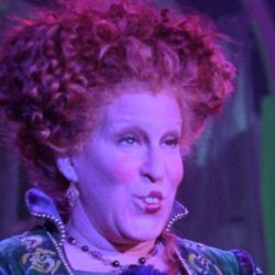 Eeb845e87a1044c5bbc88435009ea82a - bootleg betty Stage performer in a green, velvet Renaissance-style dress with a purple corset, under purple lighting, speaking or singing on stage.