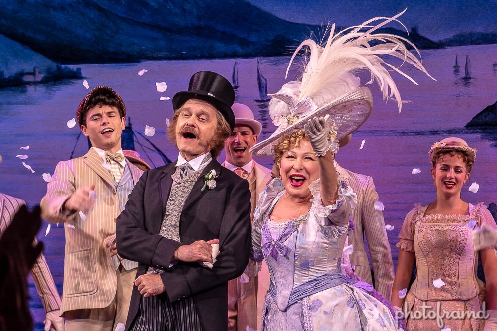 Stage performance: a man in a top hat and a woman in a silver gown with a feathered hat cheer with other costumed performers on a seaside backdrop, confetti in the air.