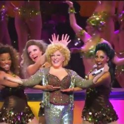 Lively stage performance with a smiling lead singer in a glittery silver outfit, flanked by backup dancers in brown sequined costumes.