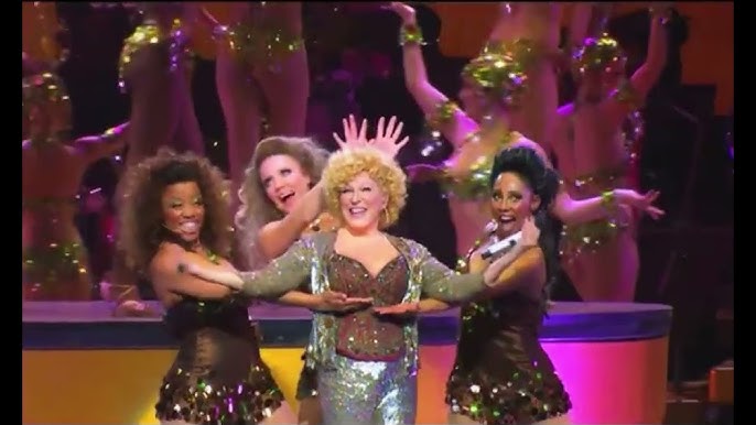 Lively stage performance with a smiling lead singer in a glittery silver outfit, flanked by backup dancers in brown sequined costumes.