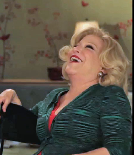 Older woman with short blonde hair laughing, wearing a green textured blazer over a red top, indoors in a warmly lit room.