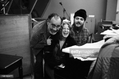 Three smiling people in a recording studio, leaning toward the camera: an older man with glasses, a woman, and a man with a turban; studio gear visible in the background.