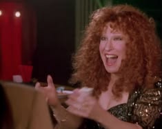 Joyful woman with curly red hair clapping and smiling, wearing a sequined top at a party or performance scene.