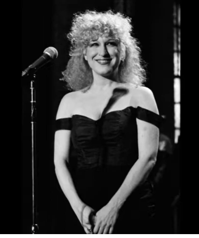 2026-08-02_11-51-05 - bootleg betty Female singer with curly blonde hair in an off-shoulder black dress on stage beside a microphone, smiling softly.