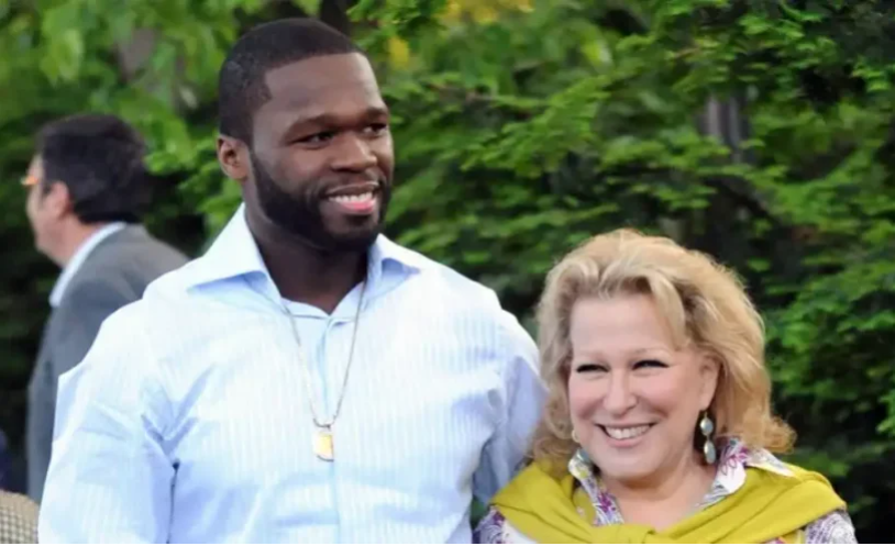 Two smiling adults posing outdoors, a man in a light blue shirt with a gold pendant and a blonde woman wearing a yellow scarf standing together.
