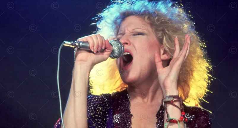 Female rock singer passionately performs into a microphone on stage, eyes closed, colorful stage lighting behind her.