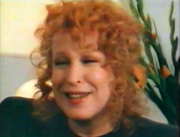 Smiling woman with curly red hair, wearing dark clothing, indoors with a tall plant in the background.