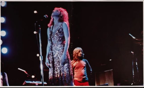 Female singer in a sequined gown performing on stage with a microphone, backed by a dancer in red pants behind her on stage.