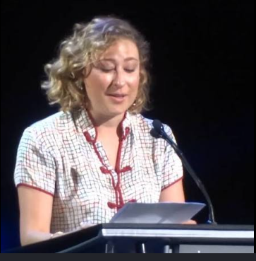 Woman with short curly blonde hair standing at a lectern, reading notes into a microphone during a speech.