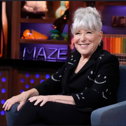 Smiling older woman with white hair, wearing a black outfit, seated in a blue armchair on a TV talk-show set with colorful shelves in the background.
