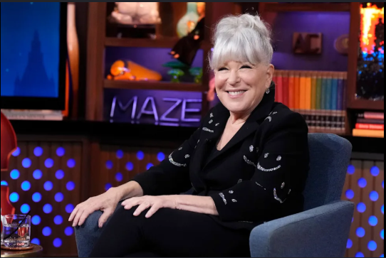 Smiling older woman with white hair, wearing a black outfit, seated in a blue armchair on a TV talk-show set with colorful shelves in the background.