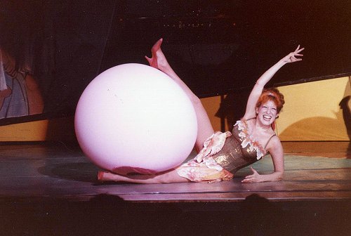A smiling cabaret dancer lying on the stage next to a large white ball prop, arms raised in a lively pose.