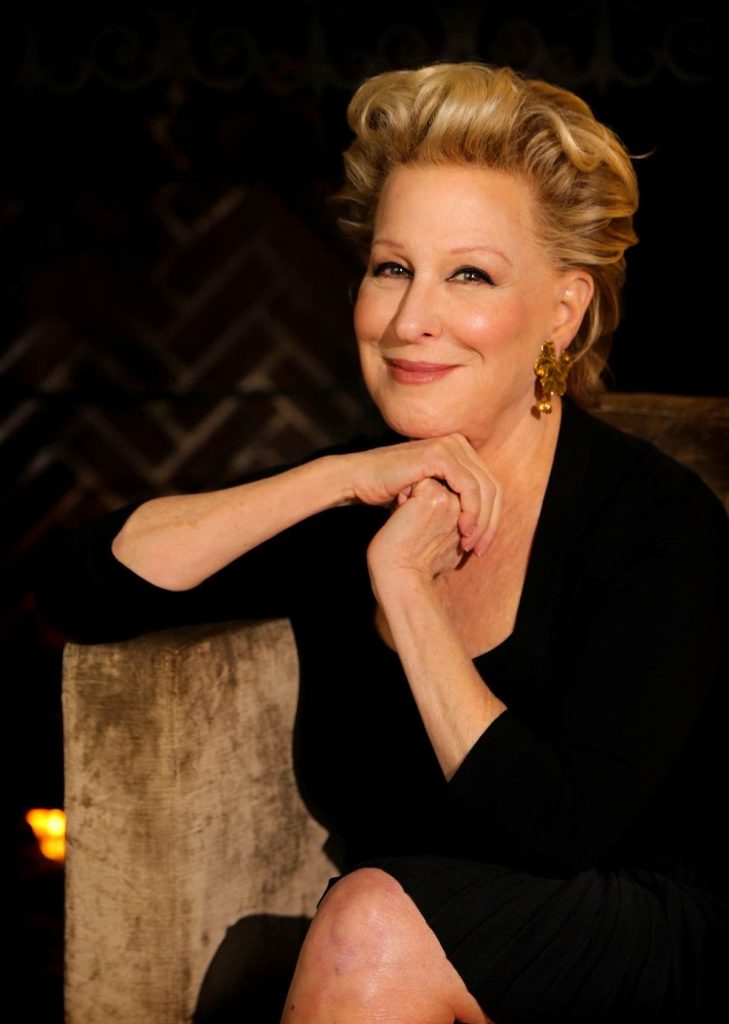 Smiling older woman with short blonde hair, wearing a black outfit and gold earrings, seated with clasped hands at her chin.