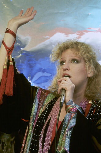 Female singer with curly blonde hair performing on stage, holding a microphone and raising her arm, wearing a colorful scarf and sparkling outfit.