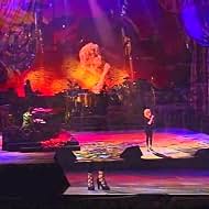 Singer performs on a brightly lit stage with a fiery lava-like backdrop and dancers in front, under purple and red lights.