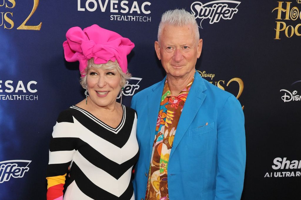 Two stylish older adults posing together on a blue carpet; woman wears a bright pink headwrap and a black?white zigzag dress, man wears a blue blazer with a tropical shirt.