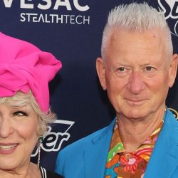 Two stylish older adults posing together on a blue carpet; woman wears a bright pink headwrap and a black?white zigzag dress, man wears a blue blazer with a tropical shirt.