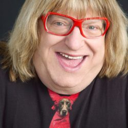 Download - bootleg betty Cheerful woman with blonde hair and red glasses, wearing a red tie with a dog graphic, against a dark background, close-up portrait