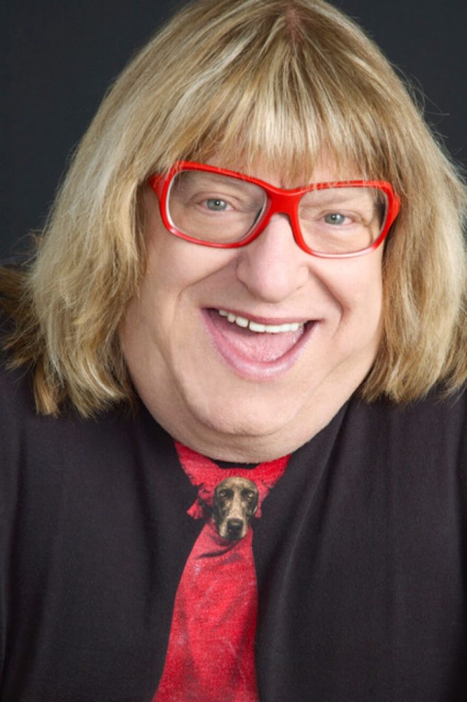 Download - bootleg betty Cheerful woman with blonde hair and red glasses, wearing a red tie with a dog graphic, against a dark background, close-up portrait