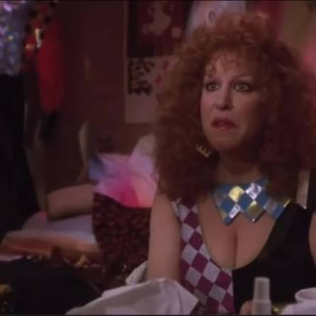 Woman with curly red hair and a surprised expression sits at a cluttered table in a dim, warmly lit room, wearing a purple-and-white diamond-pattern top and a bold diamond-shaped necklace.
