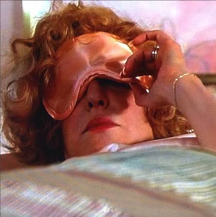 Woman lying in bed wearing a pink sleep mask being adjusted by another person on her face/eyes.