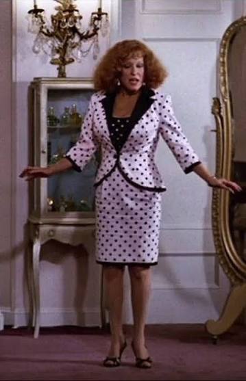 Woman with curly red hair wearing a pink polka-dot blazer, standing in a room with an antique cabinet and chandelier.
