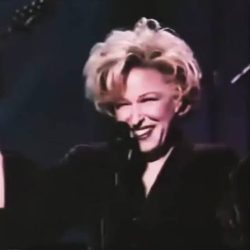 Smiling blonde female singer on stage with a microphone, arms raised, wearing a dark outfit.