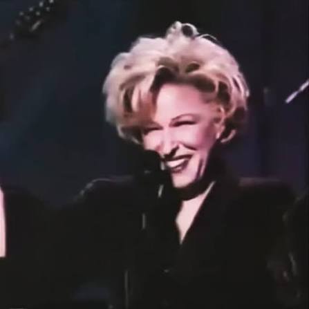 Smiling blonde female singer on stage with a microphone, arms raised, wearing a dark outfit.