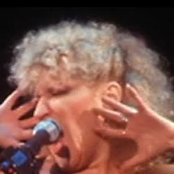 Female singer performing on stage in a gold sequined dress, hands near her face as she sings into the microphone.