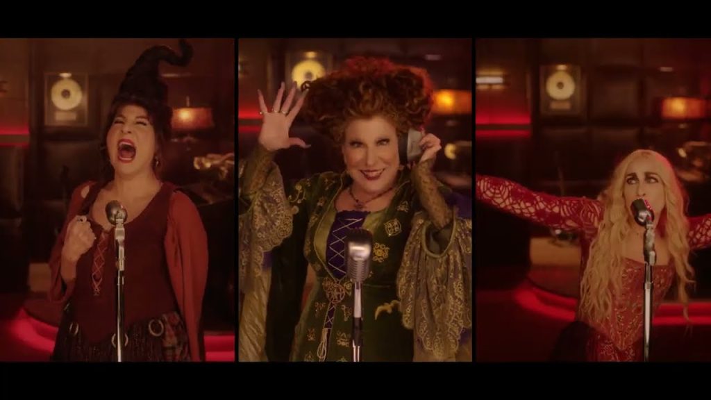 Three women in vintage costumes singing into microphones in a warm, red-lit studio.