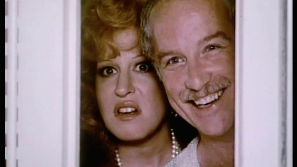 Couple peering through a doorway with shocked expressions, woman wearing a pearl necklace and a man smiling.