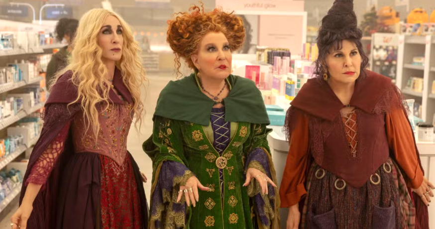 Three women in elaborate, wizard- or witch-inspired costumes stand together in a bright store aisle with shelves of products behind them.