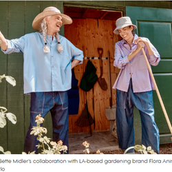 Two elderly women in wide-brimmed hats stand outside a garden shed, smiling; one leans on a rake, the other on a garden hoe with tools visible inside.